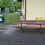 Ławka z oparciem ustawiona na nawierzchni bezpiecznej w sąsiedztwie budynku. Obok znajduje się kosz na śmieci oraz żółta skrzynka gazowa.