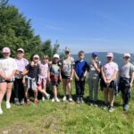 Grupa 11 dzieci stoi na tle górskiego krajobrazu z widokiem na zalesione wzgórza. Przed nimi zielona trawa, w tle bezchmurne niebo.