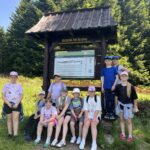 Grupa 12 dzieci pozuje pod drewnianą tablicą informacyjną Gorczańskiego Parku Narodowego, otoczoną gęstymi świerkami.