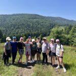 Grupa 12 dzieci wraz z dorosłą opiekunką stoi na górskim szlaku w Gorcach. W tle widać zalesione wzniesienia i niebieskie niebo.