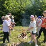 Grupa dzieci i jedna osoba dorosła pieką kiełbaski nad ogniskiem na leśnej polanie. W tle widać drzewa i plecaki leżące na trawie.