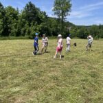 Grupa dzieci bawi się na świeżo skoszonej łące, otoczonej drzewami. W tle widać zalesione wzgórza i kilka budynków.