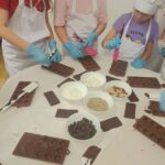 Uczennice ubrane w czapki rękawiczki i fartuchy ochronne wykonują swoje własne czekoladki w foremkach. Na środku stołu znajdują się orzechy migdały i kokos którymi dekorują swoje czekolady