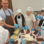 Uczniowie ubrani w czarpki, rękawiczki i fartuchy ochronne słuchają prowadzącego, ktory tłumaczy im, jak będą wykonywać swoje własne czekoladki. Uczniowie mają szpatułki i foremki.