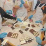 Uczniowie ubrani w czarpki, rękawiczki i fartuchy ochronne wykonują swoje własne czekoladki w foremkach. Na środku stołu znajdują się orzechy, migdały i kokos, którymi dekorują swoje czekolady.