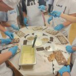 Uczniowie ubrani w czarpki, rękawiczki i fartuchy ochronne wykonują swoje własne czekoladki z białej czekolady. Na środku stołu znajdują się orzechy, migdały i kokos, którymi dekorują swoje czekolady.