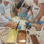 Uczniowie ubrani w czarpki, rękawiczki i fartuchy ochronne wykonują swoje własne czekoladki. Na środku stołu znajdują się pojemniki z białą i mleczną czekoladą. Uczniowie dekorują czekoladki bakaliami