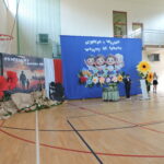 „Uroczystość rozpoczęcia roku szkolnego w sali gimnastycznej, dekoracje patriotyczne i szkolne, uczeń przy mównicy.”