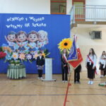 „Poczet sztandarowy podczas rozpoczęcia roku szkolnego, uczniowie w odświętnych strojach na tle dekoracji.”