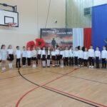 „Grupa uczniów w odświętnych strojach stoi na sali gimnastycznej podczas akademii rozpoczęcia roku szkolnego.”