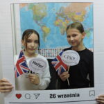Dwie dziewczyny pozują do zdjęcia w dekoracyjnej ramce z napisem Europejski Dzień Języków i datą 26 września. Obie trzymają w rękach brytyjskie flagi i napisy. W tle widoczna jest mapa świata.