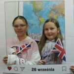 Dwie dziewczyny pozują do zdjęcia w dekoracyjnej ramce z napisem Europejski Dzień Języków i datą 26 września. Obie trzymają w rękach brytyjskie flagi. W tle widoczna jest mapa świata.