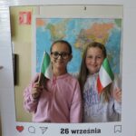 Dwie dziewczyny pozują do zdjęcia w dekoracyjnej ramce z napisem Europejski Dzień Języków i datą 26 września. Obie trzymają w rękach włoskie flagi. W tle widoczna jest mapa świata.