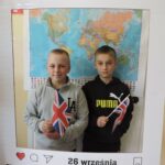 Dwóch chłopców pozuje do zdjęcia w dekoracyjnej ramce z napisem Europejski Dzień Języków i datą 26 września. Oboje trzymają w rękach brytyjskie flagi.. W tle widoczna jest mapa świata.