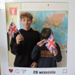 Dwóch chłopców pozuje do zdjęcia w dekoracyjnej ramce z napisem Europejski Dzień Języków i datą 26 września. Oboje trzymają w rękach brytyjskie flagi. W tle widoczna jest mapa świata.