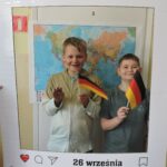 Dwóch chłopców pozuje do zdjęcia w dekoracyjnej ramce z napisem Europejski Dzień Języków i datą 26 września. Oboje trzymają w rękach niemieckie flagi. W tle widoczna jest mapa świata.