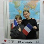 Dwoje dzieci pozuje do zdjęcia w dekoracyjnej ramce z napisem Europejski Dzień Języków i datą 26 września. Oboje trzymają w rękach francuskie flagi. W tle widoczna jest mapa świata.