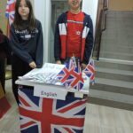 Stanowisko angielskie. Dziewczyna i chłopiec stoją za stolikiem udekorowanym flagą Wielkiej Brytanii i napisem English. Na stole rozłożone są flagi ulotki i materiały promujące język angielski.