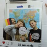 Trzy dziewczyny pozują do zdjęcia w dekoracyjnej ramce z napisem Europejski Dzień Języków. Jedna trzy ma w ręce flagę Niemiec, a dwie napisy w różnych językach. W tle widoczna jest mapa świata.