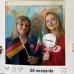 Dwie nauczycielki pozują do zdjęcia w dekoracyjnej ramce z napisem Europejski Dzień Języków i datą 26 września. Jedna trzyma w ręce niemiecką flagę, a druga napis teacher. W tle widoczna jest mapa świata.