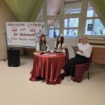 Dwie uczennice w stylizowanych strojach i nauczycielka siedzą przy okrągłym stole z czerwonym obrusem, w tle widoczna dekoracja Narodowego Czytania 2025. Na stole leżą książki i świeczniki, tworząc klimat dawnych czasów. Nauczycielka czyta na głos fragment utworu, uczennice wsłuchują się z uwagą. Wnętrze klasy ma duże okna i zielone ściany. Całość wygląda jak poetyckie spotkanie – skromne, ale pełne uroku.