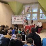 Na pierwszym planie widać uczniów siedzących na ławkach i krzesłach, zwróconych w stronę scenki. Z przodu, przy stole z czerwonym obrusem, dwie uczennice i nauczycielka czytają poezję Jana Kochanowskiego w ramach Narodowego Czytania 2025.