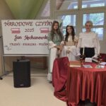 Trzy osoby — dwie uczennice w długich stylizowanych sukniach i nauczycielka w białej bluzce — stoją przed tablicą z napisem „Narodowe Czytanie 2025 Jan Kochanowski”. Obok znajduje się okrągły stół z czerwonym obrusem, świecznikami i książkami. Wnętrze klasy jest jasne i przytulne. Zdjęcie ma charakter pamiątkowy, podsumowuje wydarzenie literackie w szkole, pokazując jego uczestników w eleganckiej, nastrojowej aranżacji.