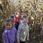 Grupa przedszkolaków spaceruje przez labirynt z wysokich łodyg kukurydzy. Dzieci są uśmiechnięte, ubrane w ciepłe ubrania, otoczone ścianą suchych roślin.