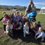 Dzieci pozują na i wokół kolorowej, niebiesko-pomarańczowej figury smoka. W tle rozciąga się górski krajobraz z boiskami i zabudowaniami. Dzieci siedzą i stoją, uśmiechając się do zdjęcia.