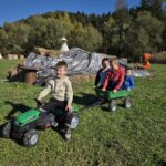 Czworo dzieci bawi się na zielonej łące, jadąc na zielonym plastikowym traktorze z przyczepką. W tle widać snopki słomy, tipi oraz zabawkowego smoka pomarańczowego koloru.