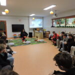 Grupa uczniów siedzi w półkolu w sali lekcyjnej i obserwuje instruktorkę pierwszej pomocy klęczącą przy fantomie do ćwiczeń resuscytacji. W tle widoczny jest ekran z prezentacją oraz ratownik w pomarańczowym stroju.