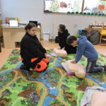 Uczniowie ćwiczą uciśnięcia klatki piersiowej na fantomach do resuscytacji, pracując pod okiem instruktorki klęczącej obok nich na kolorowym dywanie w sali lekcyjnej.