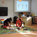 Dwóch instruktorów w strojach ratowniczych demonstruje resuscytację na fantomie; jedna osoba wykonuje uciśnięcia klatki piersiowej, a druga przygotowuje defibrylator AED, pracując na kolorowym dywanie w sali lekcyjnej.