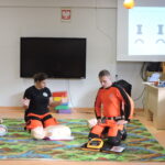 Dwóch instruktorów w strojach ratowniczych demonstruje resuscytację na Dwoje instruktorów w strojach ratowniczych demonstruje użycie defibrylatora AED na fantomie; jedna osoba unosi ręce, sygnalizując odsunięcie się od poszkodowanego, a druga klęczy obok urządzenia na kolorowym dywanie w sali lekcyjnej.