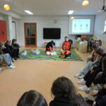 Uczniowie siedzą w dużym kręgu, obserwując dwójkę instruktorów w strojach ratowniczych, którzy demonstracyjnie wykonują resuscytację i użycie AED na fantomie leżącym na kolorowym dywanie w sali lekcyjnej.