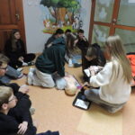 Grupa uczniów siedzi na podłodze w kąciku sali, obserwując, jak kilkoro z nich ćwiczy udzielanie pierwszej pomocy na fantomie; jedna osoba wykonuje uciśnięcia klatki piersiowej, a inne przygotowują sprzęt z apteczki pod okiem instruktora w pomarańczowym stroju.