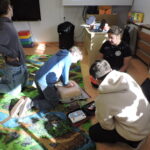 Grupa uczniów siedzi na podłodze w kąciku sali, obserwując, jak kilkoro z nich ćwiczy udzielanie pierwszej pomocy na fantomie; jedna osoba wykonuje uciśnięcia klatki piersiowej, a inne przygotowują sprzęt z apteczki pod okiem instruktora w pomarańczowym stroju.