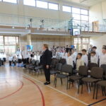 Uczniowie i nauczyciele stoją w sali gimnastycznej podczas odśpiewania hymnu.