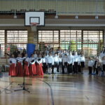 Grupa młodszych uczniów w białych koszulach i czerwonych spódnicach stoi przed mikrofonami.