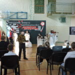 Uczniowie odgrywają scenę historyczną przed dekoracją niepodległościową, publiczność siedzi przed nimi.