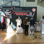 Uczniowie w strojach z różnych epok prezentują scenę patriotyczną na tle dekoracji.