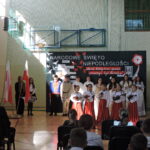 Występ skrzypaczek i grupa uczniów w strojach historycznych podczas akademii patriotycznej.