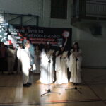 Uczniowie w białych strojach recytują przy mikrofonach podczas uroczystości szkolnej.