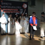 Uczniowie w kostiumach historycznych czytają tekst na scenie akademii.