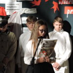 Uczennica czyta z gazety przy mikrofonie, za nią stoją uczniowie w strojach historycznych.