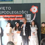 Dyrektorka przemawia do mikrofonu na tle dekoracji Święta Niepodległości, obok stoją uczniowie w kostiumach.