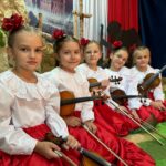 Pięć młodych dziewczynek w białych bluzkach z haftem i czerwonych, połyskujących spódnicach siedzi w rzędzie na scenie, trzymając skrzypce. Każda ma we włosach czerwoną kwiatową ozdobę. W tle znajdują się biało-czerwone tkaniny, czarna sylwetka powstańca oraz duży plakat z wizerunkiem Zamku Królewskiego, tworzące patriotyczną scenografię