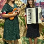 „Dwie nastoletnie dziewczyny stoją na tle patriotycznej dekoracji przedstawiającej Zamek Królewski i sceny powstańcze. Ubrane w ciemne, grochowe sukienki, mają na ramionach biało-czerwone opaski. Jedna trzyma skrzypce, druga akordeon. Pod ich stopami leży zielona i brązowa tkanina stylizowana na sceniczne podłoże