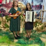 „Dwie nastoletnie dziewczyny stoją na tle patriotycznej dekoracji przedstawiającej Zamek Królewski i sceny powstańcze. Ubrane w ciemne, grochowe sukienki, mają na ramionach biało-czerwone opaski. Jedna trzyma skrzypce, druga akordeon. Pod ich stopami leży zielona i brązowa tkanina stylizowana na sceniczne podłoże