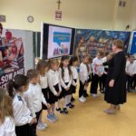 Pierwszoklasiści stoją w rzędzie podczas uroczystego pasowania, wysłuchując słów nauczyciela